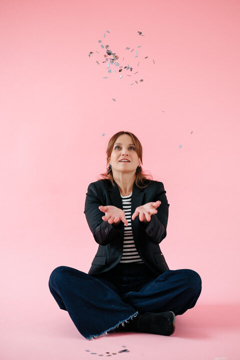 Nicole Forrai sitzt in einem rosafarbenen Raum und wirft Konfetti in die Luft, das symbolisch für die vielen Kompetenz einer Person steht, die in der Kompetenzbilanz analysiert wurden.