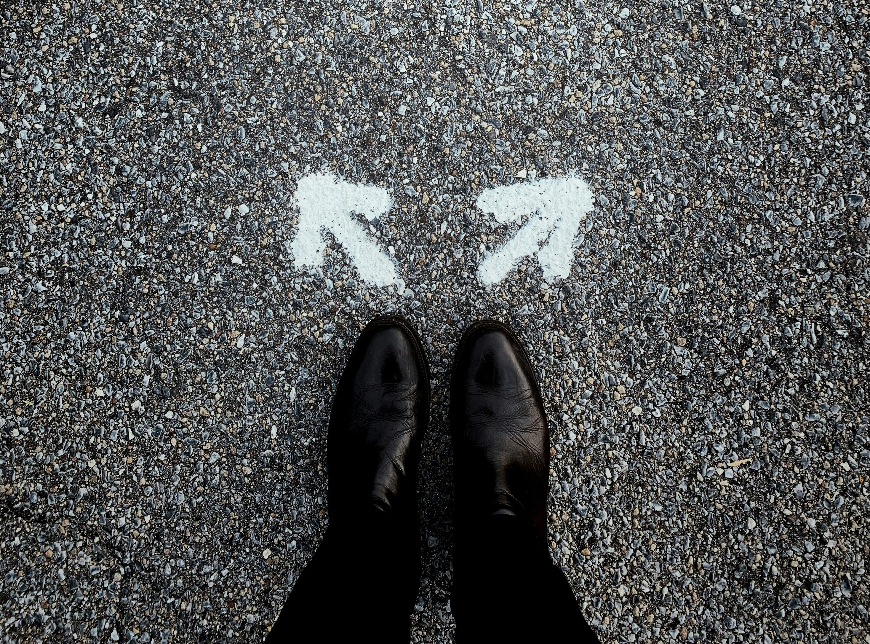 Das Bild zeigt Füße in Schuhen auf einer asphaltierten Straße. Vor den Schuhen sind zwei Pfeile die in entgegengesetzt Richtungen zeigen, sie stehen für einen Entscheidungsweg im Karriere Coaching.