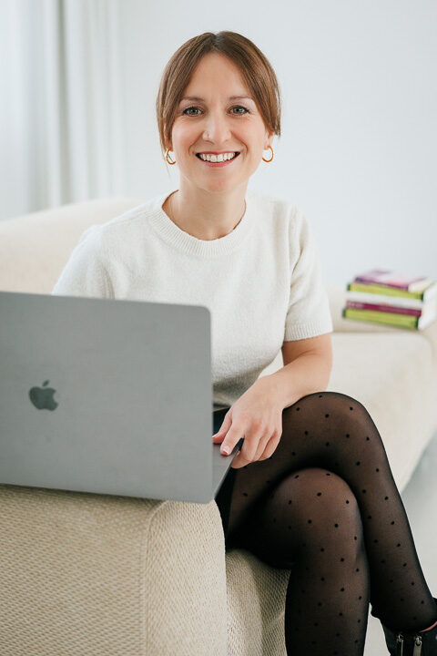 Nicole Forrai sitzt auf einem weißen Sofa. Auf der Lehne steht ein Laptop, den sie für Online-Coachings nutzt.